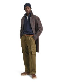 Bruin geruite jas, marineblauwe rits-trui, groene cargobroek, bruine gevlochten riem en bruine schoenen met witte zolen. Wolstof met zakken.