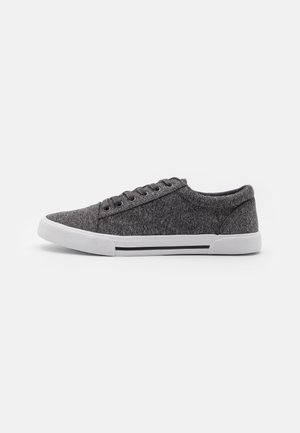 Zapatilla baja de tela gris con ojales negros, suela de goma blanca y detalle de raya negra sobre fondo blanco liso.