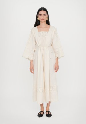 Cream linen dress with a square neckline, wide sleeves, and eyelet lace detailing. Drawstring waist and ankle-length silhouette. Black shoes.
