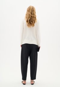 Donna con lungo capelli biondi mossi, che indossa un maglione bianco, pantaloni neri a vita alta, sandali neri, mentre tiene una pochette nera, rivolta di spalle.