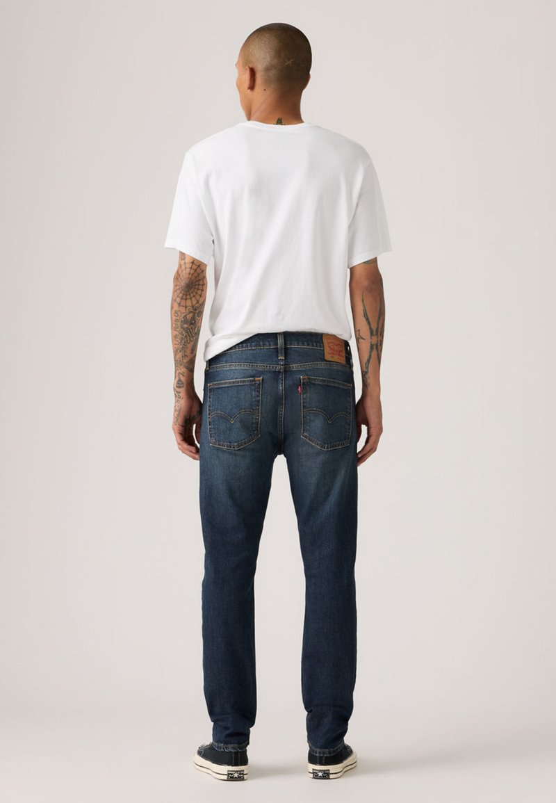 Jean en denim bleu foncé avec une coupe droite, doté de poches arrière et d'un patch en cuir de la marque. Associé à un t-shirt blanc uni.