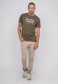 T-shirt vert olive avec le texte blanc "BLUE&JEANS", associé à un pantalon beige clair. Le mannequin porte des baskets blanches et se tient avec une main dans la poche.