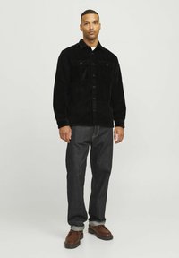 Black corduroy shirt jacket with two chest pockets, button closure, worn over a white shirt, paired with dark denim jeans and brown shoes.