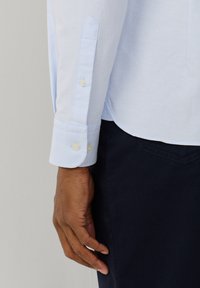 Mano dalla pelle scura appoggiata accanto al corpo, con polsino di camicia a manica lunga azzurro chiaro e pantaloni blu scuro.