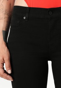 Černé džíny s vysokým pasem, které mají přední kapsy, knoflíkový zip a hladkou texturu. Jsou viditelné jemné detaily prošití.