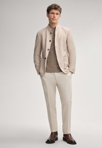 Beige Jacke mit Stehkragen, braunem Pullover, hellen Hosen und braunen Knöchelstiefeln. Weicher Stoff und Knopfdetails an der Jacke.