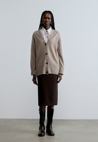 Helepruun kootud kardin, millel on suured nööbid, kantud valge triiksärgi ja lipsuga, kombineeritud pruuni midi seeliku ning põlveni ulatuvate mustade saapadega.