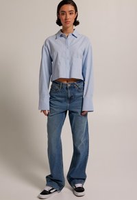 Light blue cropped button-up shirt with a single chest pocket, paired with loose-fitting, mid-blue jeans and black sneakers.