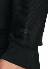 Lähikuva mustast vatiinivaatetehnoloogiategija käistest, millel on ribbiga mansett ja väike must Under Armouri logo alumisel käeosal.