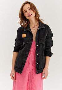 Czarna kurtka dżinsowa z guzikami i dwoma przednimi kieszeniami, nałożona na różową sukienkę z falbankami i dekoltem w kształcie litery V.