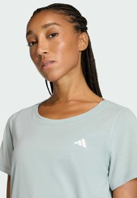 Žena s pletenými vlasmi, oblečená v svetlomodrom tričku s malým bielym logom Adidas na hrudi, na jednoduchom pozadí.