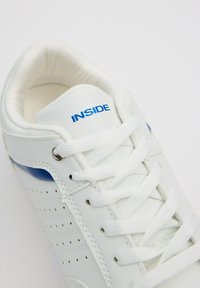 Zapatilla blanca con detalles azules, de piel suave, parte superior perforada, cordones planos y cuello acolchado. Logo "INSIDE" impreso en azul en la lengüeta.