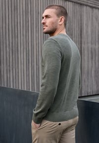 Homme aux cheveux courts portant un pull vert et un pantalon beige, debout les mains dans les poches, regardant sur le côté, devant un mur en bois et métal.