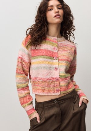 Kobieta o falujących brązowych włosach ma na sobie krótki, wielokolorowy, w paski kardigan z różowymi guzikami oraz brązowe spodnie z wysokim stanem, ręce w kieszeniach.