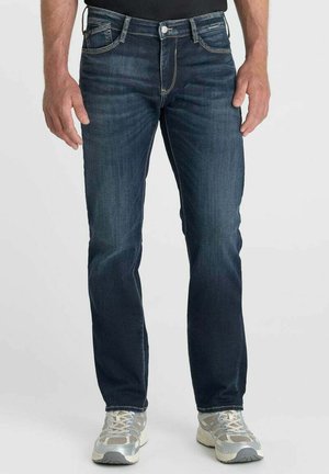 Man draagt donkerblauwe slim-fit jeans en grijze sportschoenen en staat voor een effen achtergrond.