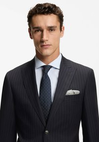 Granatowa marynarka w prążki z klapami z wycięciem, biała koszula w niebieskie paski, krawat w groszki oraz szara poszetka widoczna w kieszeni marynarki.