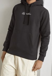 Felpa nera con cappuccio, dotata di tasca frontale, caratterizzata da un logo ricamato bianco. Tessuto morbido, polsini e orlo a costine, vestibilità comoda. Indossata con pantaloni di colore chiaro.