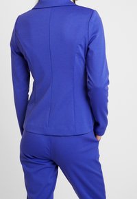 Veste ajustée bleue à manches longues, avec une texture douce et un col classique. Présente des détails de coutures le long du dos et des poches.