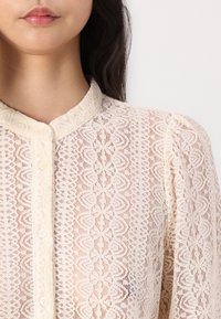 Blusa de encaje en suave beige con cuello alto y cierre de botones. Presenta intrincados patrones florales y una delicada textura semitransparente.