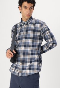 Ναυτικό καρό πουκάμισο με κουμπιά σε σκούρο μπλε, με ανοιχτό μπλε, λευκές και μπορντώ αποχρώσεις. Διαθέτει τσέπη και καμπυλωτό τελείωμα. Το μοντέλο κρατά μια μαύρη τσάντα.