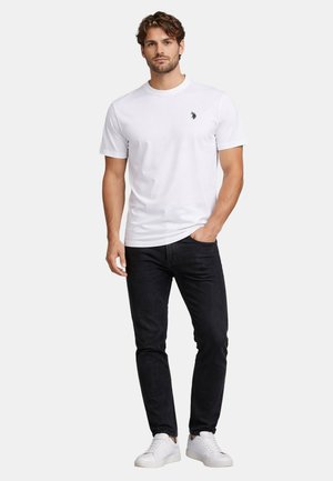 Mann trägt weißes T-Shirt mit kurzen Ärmeln und kleinem schwarzem Logo, schwarze Jeans und weiße Sneaker, steht mit einer Hand in der Tasche.