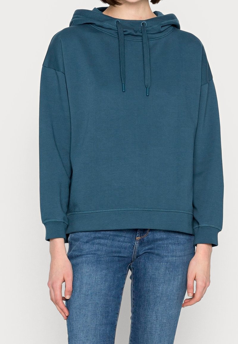 Personne portant un sweat à capuche bleu sarcelle avec des cordons et un jean bleu, debout devant un fond clair uni.