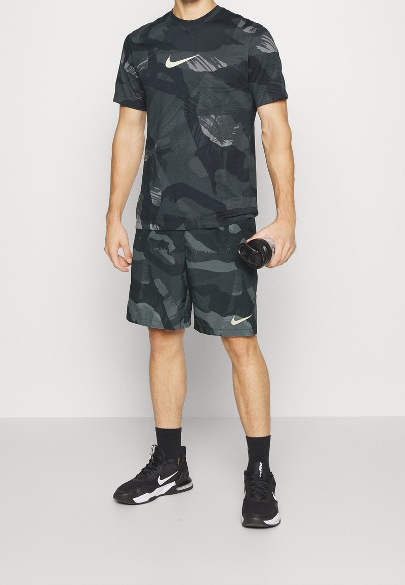 Homme portant un t-shirt et un short de sport Nike camouflage noir et gris, baskets noires, tenant une bouteille d'eau noire.