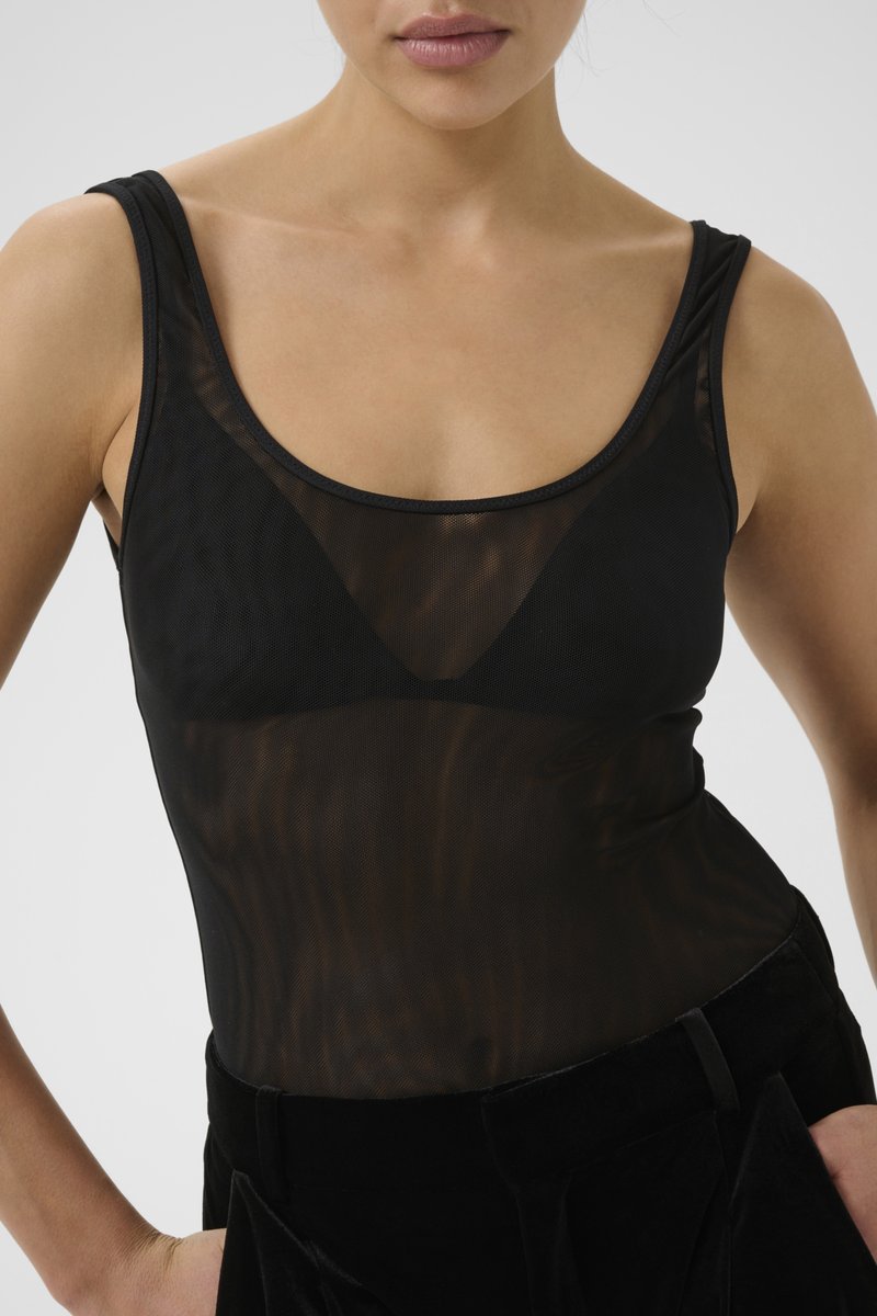 Woman wearing a black sheer sleeveless top over a black bra, paired with black pants, arms bent with hands on hips against a plain background.