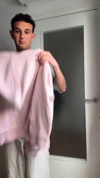 Jeune homme aux cheveux courts et foncés tenant un pull rose clair dans une pièce avec une porte blanche fermée avec du verre dépoli.