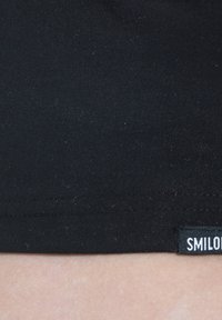 Sort stof i nærbillede af en skjorte underkant med en lille mærkat med teksten "SMIL." Glat tekstur med en tætsiddende form. Ingen mønstre eller accenter synlige.