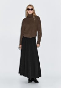 Maglione a collo alto a coste marrone e gonna maxi nera con bottoni. Il modello indossa occhiali da sole neri e stivali alla caviglia. Texture liscia, vestibilità su misura.