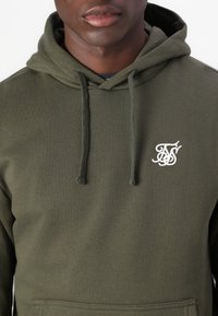 Sudadera verde oliva hecha de una tela suave, con un bolsillo frontal y cordones. Presenta un logo bordado en blanco en el pecho.