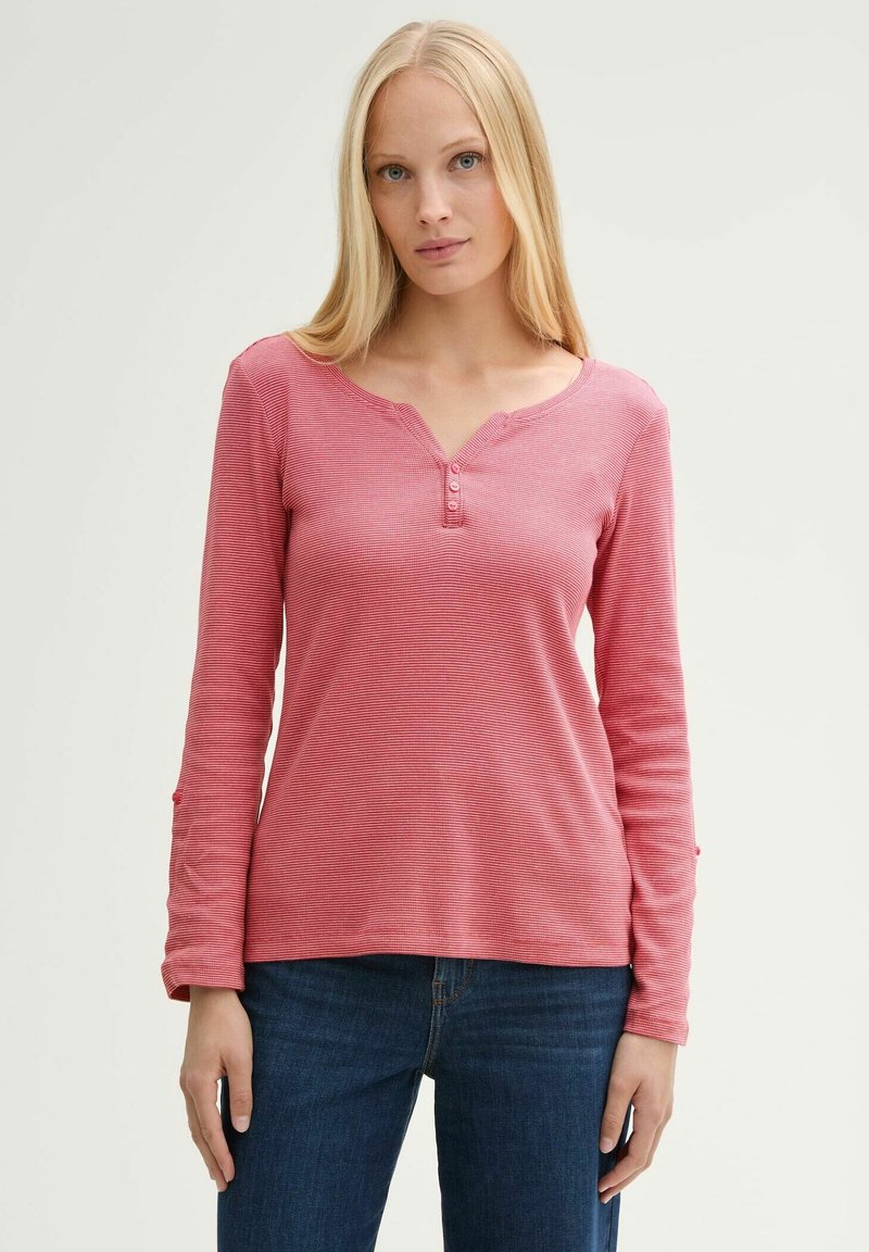 Rosa Langarmshirt mit Henley-Ausschnitt, das drei Knöpfe und eine gerippte Textur aufweist. Kombiniert mit blauen Jeans.