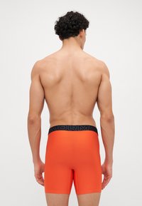 Oransje boxertruser med en tettsittende passform og svart midjebånd som viser ordet "MORMOR." Stoffet ser glatt og elastisk ut.