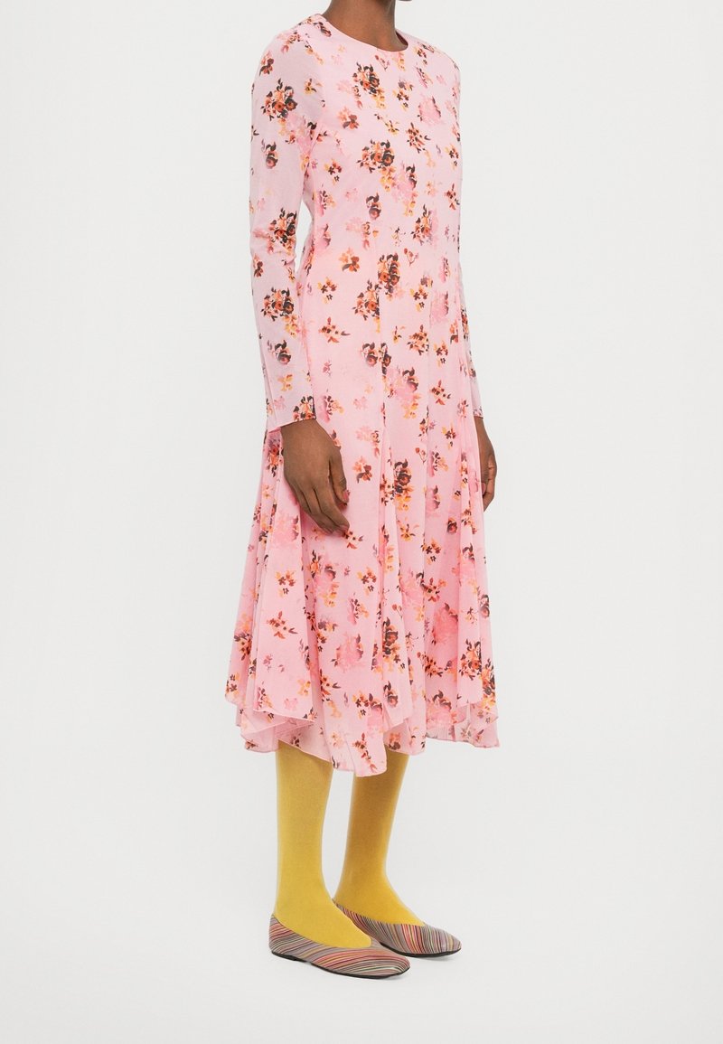 Femme portant une robe fleurie rose à manches longues, des collants jaunes et des chaussures plates rayées multicolores, debout sur un fond uni.