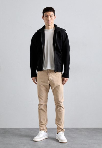 Zwarte gebreide zip-up jas over een wit shirt, gecombineerd met beige broeken en witte sneakers, staande tegen een neutrale achtergrond.