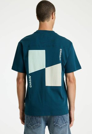 Man draagt een teal t-shirt met twee grote witte geometrische vormen en de tekst "CHASIN'" op de achterkant, gecombineerd met een blauwe spijkerbroek.
