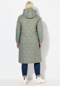 Lange, met een kap, olijfgroene gewatteerde jas met een ruitpatroon, mouwen van volledige lengte en zijzakken, gecombineerd met blauwe spijkerbroeken en witte laarzen.