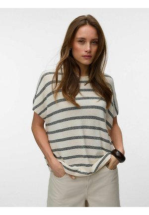 Femme aux longs cheveux bruns portant un haut à manches courtes crème et noir à rayures, un pantalon beige et un bracelet foncé, mains dans les poches.