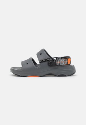 Mørkegrå Crocs-sandal med to åbne stropper, en aftagelig mønstret ankelrem og orange accenter på en hvid baggrund.