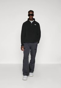 Vyras, vilkintis juodą „North Face“ puloverį, tamsiai pilkas kelnes su užtrauktukais kišenėse, baltus sportinius batelius ir juodus saulės akinius, stovintis prieš paprastą foną.