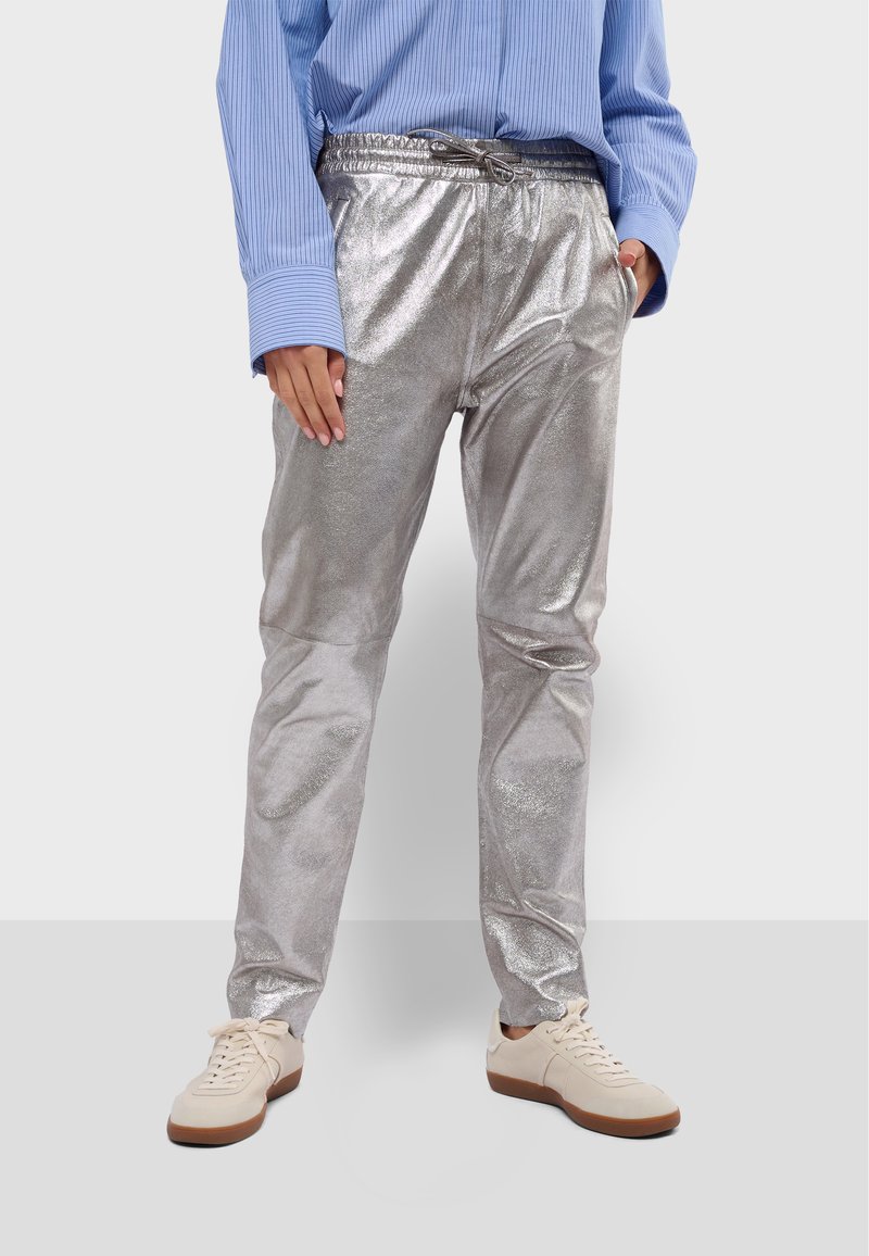 Zilveren metallic broek met een glanzende afwerking, elastische tailleband en trekkoord. G gedragen met een lichtblauw gestreept shirt en beige sneakers.
