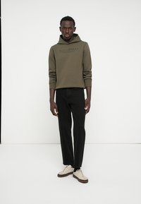 Homme se tenant contre un fond blanc, portant un sweat à capuche vert olive, un pantalon noir et des baskets beiges avec des semelles marron.