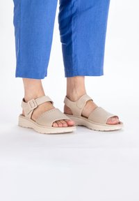 Beige sandals with a woven texture, adjustable buckle, and a cushioned sole. Paired with cropped blue pants, showcasing open toes.