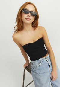 Zwarte geribbelde tube-top gecombineerd met lichtblauwe high-waisted jeans. Het model draagt donkere kattenoog-zonnebrillen en heeft golvend rood haar.