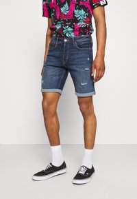 Mies, joka pukeutuu tummansiniset käärittyihin farkkushortseihin, valkoisiin sukkiin, mustiin lenkkareihin ja kirkkaasti kukkakuosisesta lyhythihaisesta paidasta, seisoo yksinkertaista taustaa vasten.