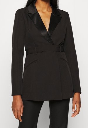 Vrouw die een zwart blazer met ceintuur en satijnen revers draagt en bijpassende zwarte broek, op een effen lichte achtergrond.