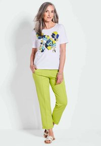 Weißes T-Shirt mit blauem und gelbem Blumenmuster; kombiniert mit grünen Hosen mit Stichdetails; Modell trägt beigefarbene Sandalen.