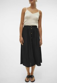 Gonna midi nera con vita arricciata e patta frontale con bottoni. Abbinata a una canotta bianca a costine e sandali neri slip-on.