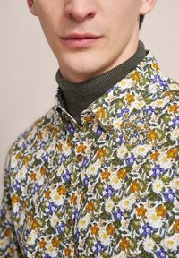 Bluse mit Blumenmuster auf weißem Grund, die orangefarbene, gelbe und lila Blumen zeigt. Braune Knöpfe und ein grüner strukturierter Rollkragen darunter.